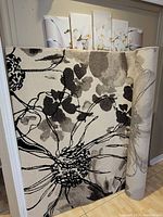 Partially unrolled rectangular area rug leaning against wall on hardwood floor showing large abstract floral black and gray design on off-white background