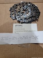 Photo of metal chainsaw chain coiled on cardboard background with paper showing chain specifications (25", 84T, 0.50 gauge, 3/8 pitch) and Stihl informational printout.