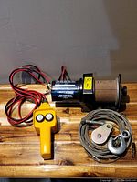 Photo showing the Centric branded 0.5 HP 12V electric winch with attached wiring, yellow wired remote control, and coiled steel cable with hook and pulley.