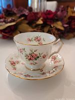 Teacup and saucer set with visible rose cluster pattern, gold trim, and white background. The set is arranged on a white surface with a floral backdrop.