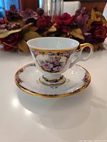 Tea cup and saucer set with visible detailed rose patterns and heavy gold trimming, photographed on a white tabletop with decorative background.