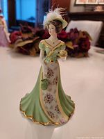 Front view of the detailed porcelain bell showing the lady holding a cup of tea and handbag in a green dress with floral patterns and gold trim.