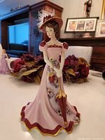 Front angle photo of the Bradford Editions porcelain lady figurine showing detailed pink gown with floral pattern, maroon hat with feathers, and umbrella with gold trim.