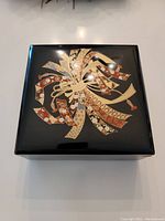 Top view showing black lacquer box with lid decorated by intricate multi-colored ribbon pattern in gold, black, white, and red.