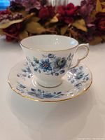 View of bone china footed tea cup and saucer with blue rose floral pattern and gold rim against floral background.