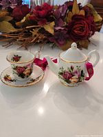 Teapot and teacup with saucer ceramic ornaments decorated with floral rose pattern and gold trim, placed on white surface with floral backdrop.