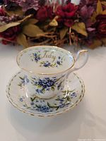 Teacup and saucer set with floral Forget Me Not pattern and inscribed "July" on cup's inner rim.