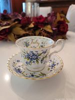 Teacup and saucer showing overall pattern with blue forget me not flowers, 'July' marking, and gold accents