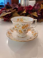 Teacup and saucer displayed with floral background, showing the yellow flower and green leaf design, gold trim on edges, and ornate handle on the teacup.