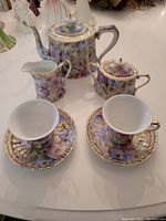 Photo shows full tea set with teapot, creamer, sugar bowl with lids, two footed teacups and two saucers decorated with pastel blue, pink, and purple morning glory flowers and gold gild accents.