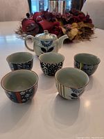 Photo shows the full set of one teapot and five cups placed on a white surface with floral decoration in background. Teapot has a lid and painted blue floral pattern.