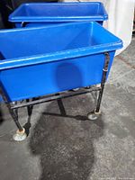 Front-side angle of blue plastic basket truck on black metal frame with white caster wheels, showing basket interior and frame condition.