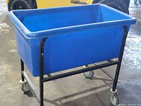 Front angled view of a blue plastic basket truck with black metal frame and four caster wheels standing on concrete floor.
