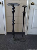 Two tall solid brass candle holders standing on floor, one taller and slimmer, both with round bases and spikes on top.