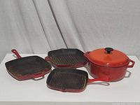 Three red-orange enamel cast iron griddle skillets and one dutch oven with lid arranged on white surface against white backdrop.