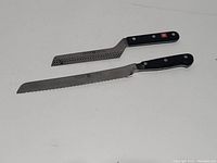 Two Wusthof serrated knives on a white surface, one with intact black handle and one with damaged handle piece.
