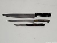Three kitchen cutting tools arranged horizontally on a white surface: a large chef's knife, a long narrow knife, and a small serrated knife. All have signs of use and require cleaning.