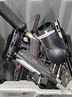 Box containing multiple paintball markers with attached air canisters, black hoppers, masks, and accessories all mixed together.