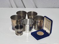 Photo of 4 tarnished silverplate KFC cups, 1 silverplate Olympic desk cup, and a Medal of Merit coin in display box