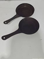 Top-down view of two small cast steel camp skillets on white surface; handles aligned to the left with hanging holes visible. Both skillets show seasoned patina. One skillet handle is warped slightly.