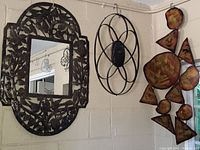Three wall hangers displayed on a beige wall: a brown carved wooden mirror with bird and tree motifs on the left, a black metal circular wall art with an oval center in the middle, and an orange abstract metal wall art composed of various circular and triangular shapes on the right.