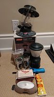 Coleman propane lantern on box with two Coleman battery lanterns in packaging, small battery lantern, blue BBQ lighter, matchbook, fire paste tube, and a plastic candle lantern.