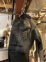 Black Batman costume with muscle padding and hood hanging in a basement area showing muscle texture and design.