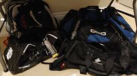 Four bags arranged on floor: large Ogio 6900 duffel, Under Armour Maple Leafs duffel, Eddie Bauer and Targus backpacks visible.