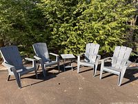 Four gray plastic Adirondack chairs arranged around one gray plastic folding table outdoors on pavement with greenery in the background.