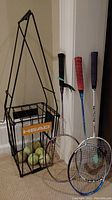 Full view of Head branded black metal tennis ball basket with multiple tennis balls inside and three badminton rackets leaning against wall, showing different colored grips
