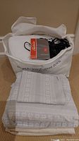 Heated mattress pad inside white fabric storage bag labeled 'European White Goose Down'. Sunbeam manual and black controller visible on top.