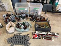 Wide shot showing two plastic bins with loose LEGO parts, partially assembled castle sections, minifigure box, track rails, fire truck and train cars