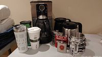 Wide photo showing the Oster coffee maker, glass carafe, two Keurig carafes, and multiple travel mugs and flasks including Terminator cup and emergency flask.