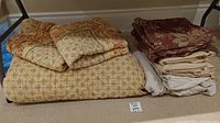 View of the folded yellow quilted coverlet and pillow shams with floral and geometric patterns, next to folded brown and beige bed linens.
