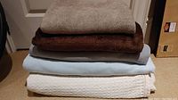 Stack of five throw blankets showing different colors and textures including white waffle pattern, light blue fleece, gray polyester, brown fleece and beige knit.