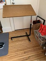 Bed table with wood-grain rectangular top angled upward on a black metal frame with wheels visible.