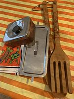 Set of kitchen items on orange and white striped surface: tiki wooden spoon and fork, two metal canisters with lids, and tea towel with vegetable design.