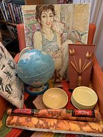 Photo shows world globe, original painting of a woman, stacked Melmac plates, McCoy bowls, wooden tulip wall hanging, two rolls of floral contact paper, and a small wooden nose-shaped glasses holder arranged on a red chair with crocheted cover.