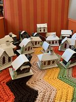 Overview of 11 Putz style paper houses and churches on a multicolored textile surface. The houses have small brush trees and holes in the back for Christmas lights.