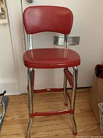 Front view of red vinyl cushioned stool with chrome legs and footrest bars, showing the seat and backrest.
