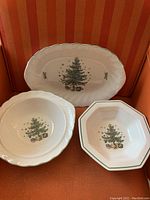 Three ceramic serving pieces, one platter and two bowls, all decorated with Christmas tree design and embossed edges, shown on orange chair