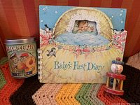 Photo showing the Vintage Baby's First Diary book with colorful baby and bird illustration, Peach-ee Keen tin for baby bib, all placed on a multicolor crocheted blanket.