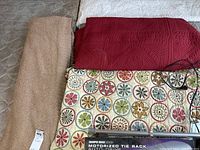 Four blankets and quilts laid out on a bed: beige blanket, red quilt, white patterned quilt, multicolored floral blanket.