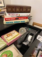 Stack of books including the complete works of Shakespeare, No Easy Day, Killing Kennedy, and Practical Problem Solver with office supplies including stapler, tape dispenser, calculators, paperweight, and box of thank you cards