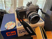 Nikon Nikkormat FTN camera with Nikon branded strap on a box of Sonomor Super Steel safety pins, round Pikachu pin next to camera, all on table with lace curtain background.
