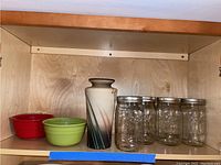 Image showing two ceramic bowls, one red and one green, alongside a tall ceramic vase and seven Ball canning jars with lids on a wooden shelf.