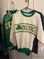 White knit Celtic sweater and green satin Celtics jacket hanging side by side with dark green Irish True T-shirt behind