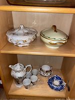 Shelf with two lidded serving dishes on top, teapot, four teacups, two teacups with saucers, and one blue and white teacup with saucer and spoon below