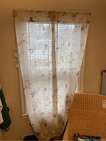 Curtain panels hung on window showing semi-transparent floral white fabric with pink and purple flowers. Light passing through reveals fabric details and room background.