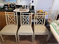 Four wood dining chairs with beige upholstered seats placed indoors near kitchen counter, showing natural wear and fabric texture